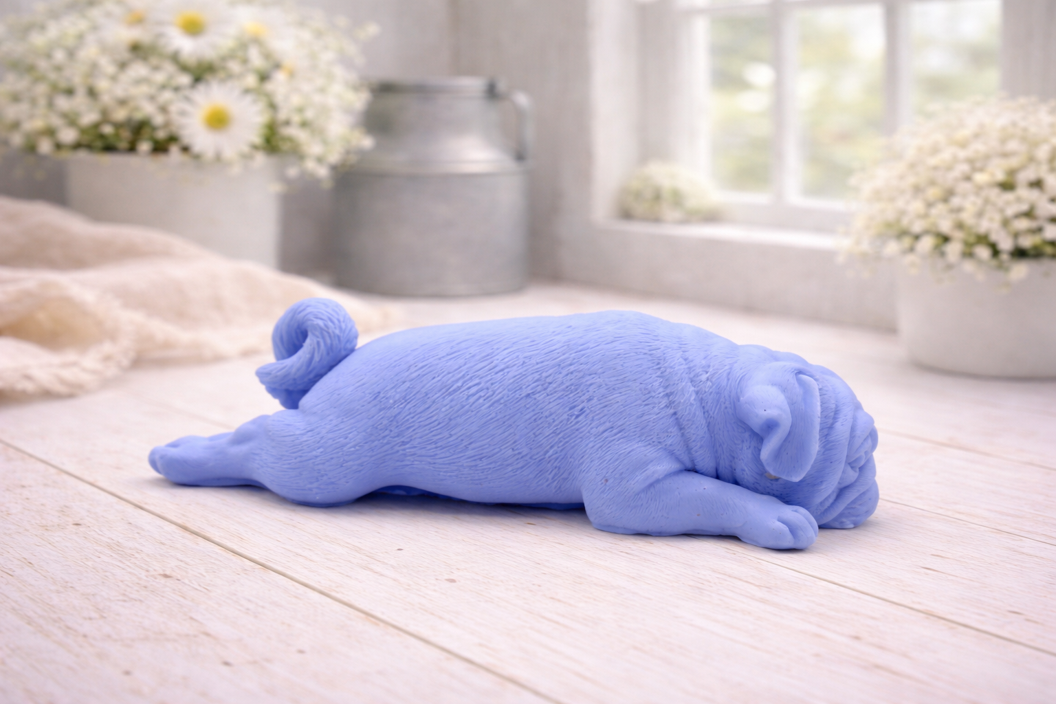 A detailed handmade vegan soap gift, sculpted in the shape of a pug dog lying flat on its stomach in a sleeping position, showing realistic skin folds and fur texture, displayed on a light wooden surface. Handmade in Blackpool 