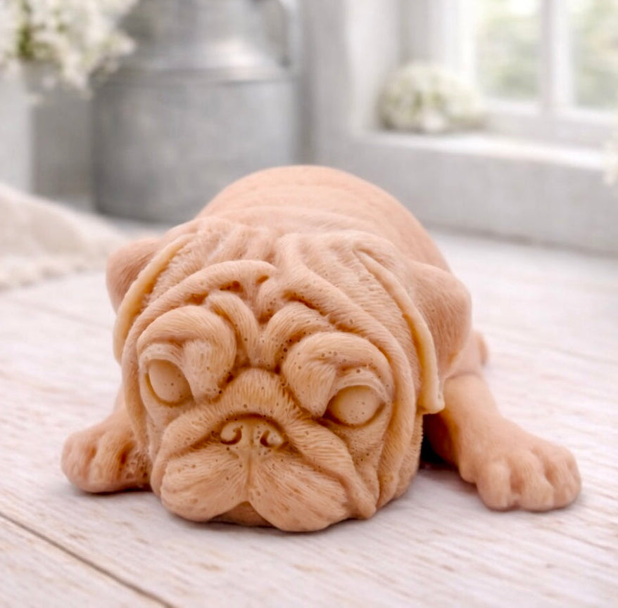 A detailed handmade vegan soap gift, sculpted in the shape of a pug dog lying flat on its stomach in a sleeping position, showing realistic skin folds and fur texture, displayed on a light wooden surface. Handmade in Blackpool 