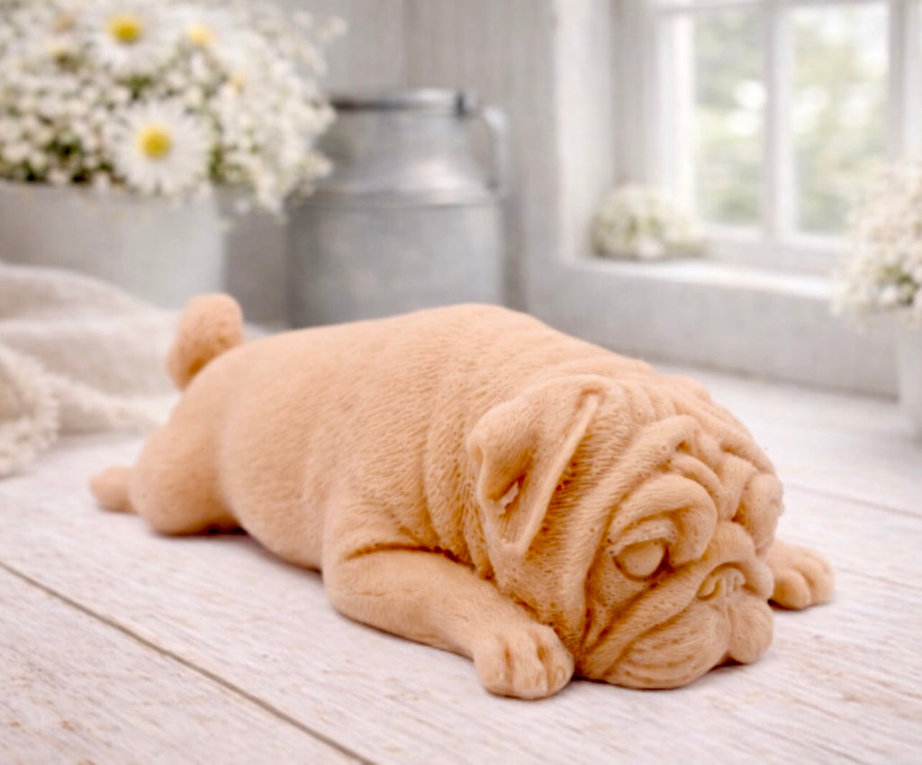 A detailed handmade vegan soap gift, sculpted in the shape of a pug dog lying flat on its stomach in a sleeping position, showing realistic skin folds and fur texture, displayed on a light wooden surface. Handmade in Blackpool 