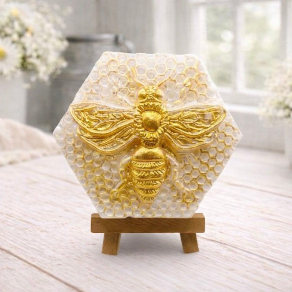 A hexagonal handmade vegan soap featuring a detailed relief of a large queen bee with outstretched wings, highlighted in shimmering gold, set against a textured white honeycomb background and displayed on a miniature wooden easel.