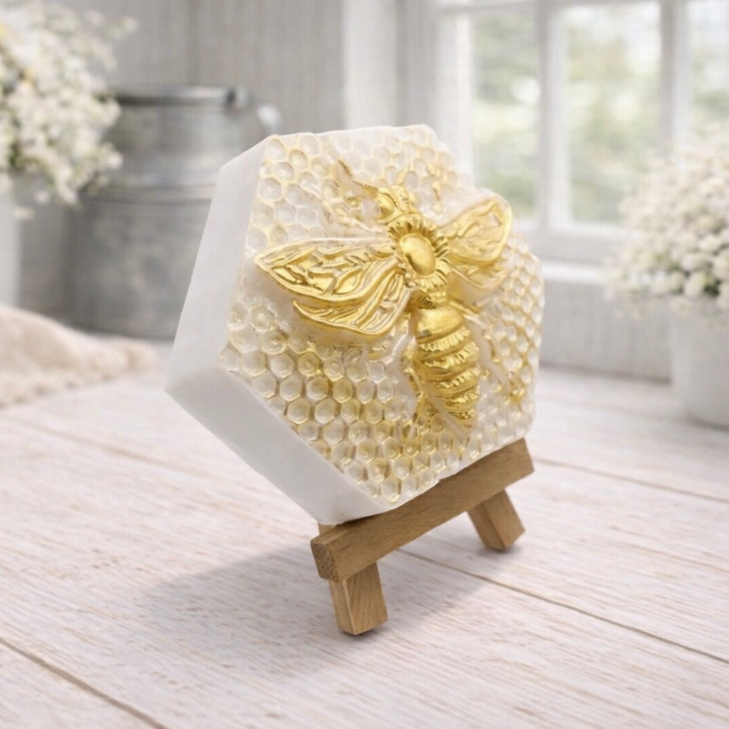 A hexagonal handmade vegan soap featuring a detailed relief of a large queen bee with outstretched wings, highlighted in shimmering gold, set against a textured white honeycomb background and displayed on a miniature wooden easel.