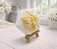 Load image into Gallery viewer, A hexagonal handmade vegan soap featuring a detailed relief of a large queen bee with outstretched wings, highlighted in shimmering gold, set against a textured white honeycomb background and displayed on a miniature wooden easel.
