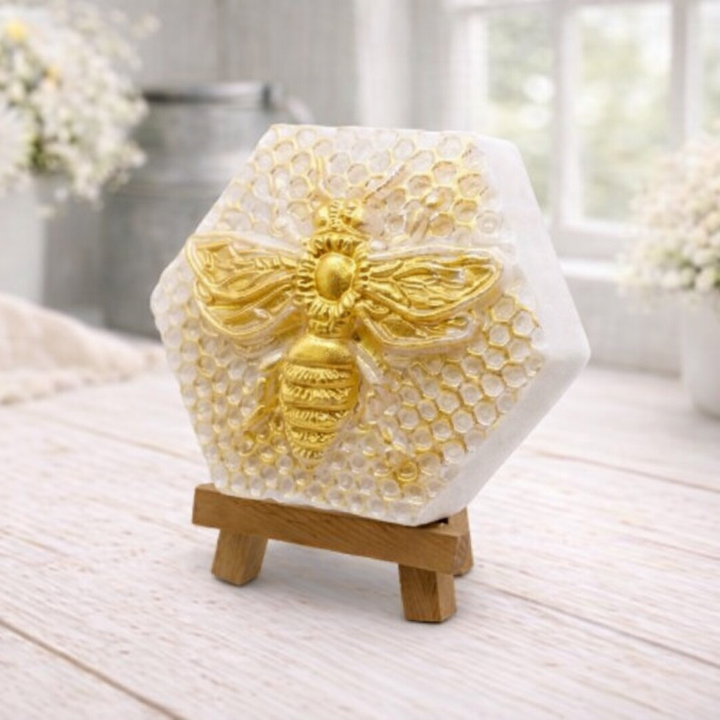 A hexagonal handmade vegan soap featuring a detailed relief of a large queen bee with outstretched wings, highlighted in shimmering gold, set against a textured white honeycomb background and displayed on a miniature wooden easel.