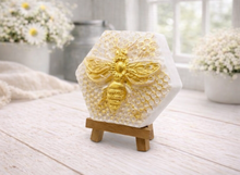 Load image into Gallery viewer, A hexagonal handmade vegan soap featuring a detailed relief of a large queen bee with outstretched wings, highlighted in shimmering gold, set against a textured white honeycomb background and displayed on a miniature wooden easel.
