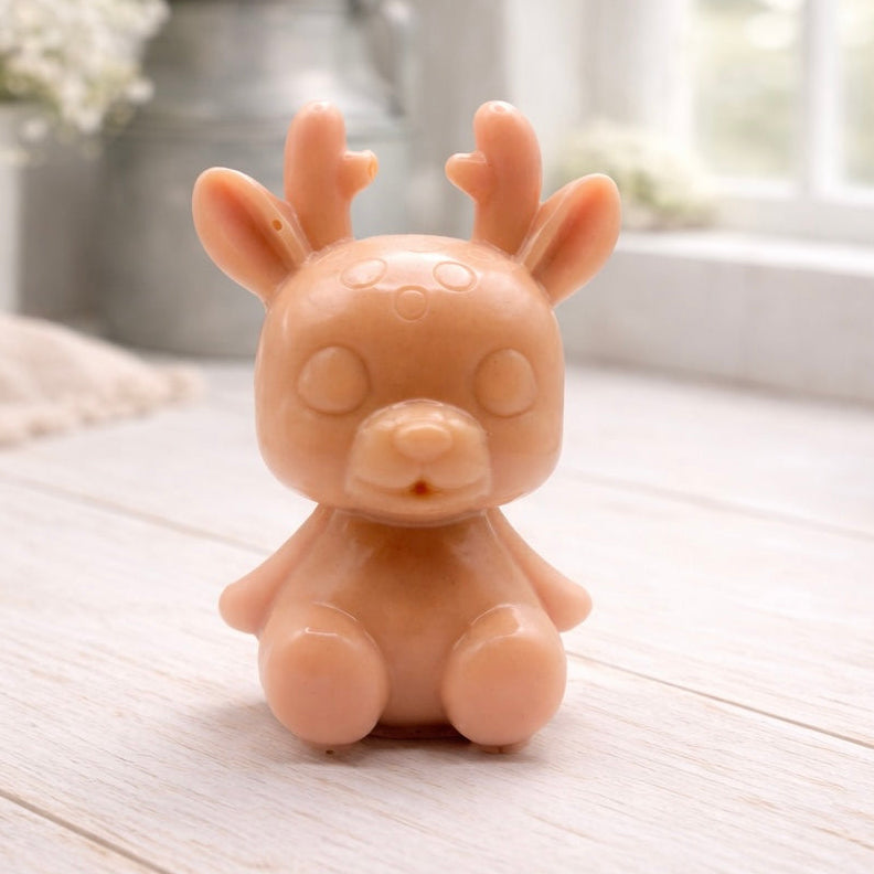 A 3D handmade vegan soap crafted into a cute, sitting baby reindeer with small antlers, rounded features, and a sweet expression, set against a bright farmhouse window.