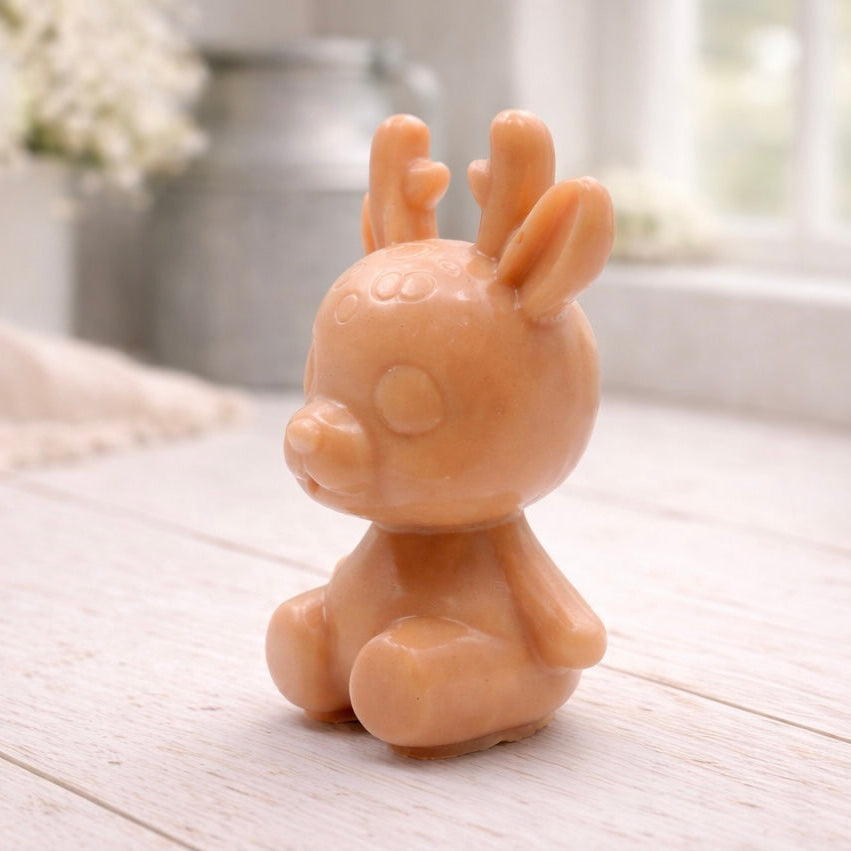 A 3D handmade vegan soap crafted into a cute, sitting baby reindeer with small antlers, rounded features, and a sweet expression, set against a bright farmhouse window.