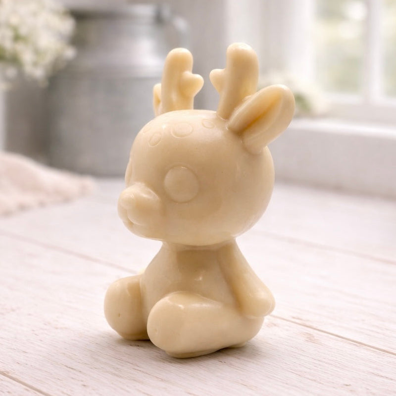 A 3D handmade vegan soap crafted into a cute, sitting baby reindeer with small antlers, rounded features, and a sweet expression, set against a bright farmhouse window.