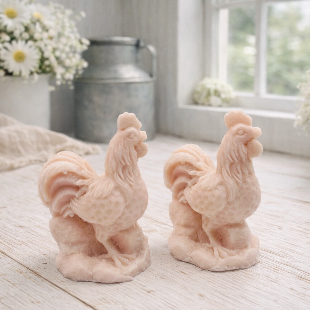 Two handmade vegan soaps sculpted in the shape of proud roosters (cockerels) with detailed feathers, combs, and curved tail feathers, standing on a rustic base, in a natural colour. Gift boxed 