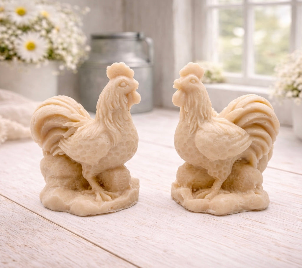 Two handmade vegan soaps sculpted in the shape of proud roosters (cockerels) with detailed feathers, combs, and curved tail feathers, standing on a rustic base, in a natural colour. Gift boxed 