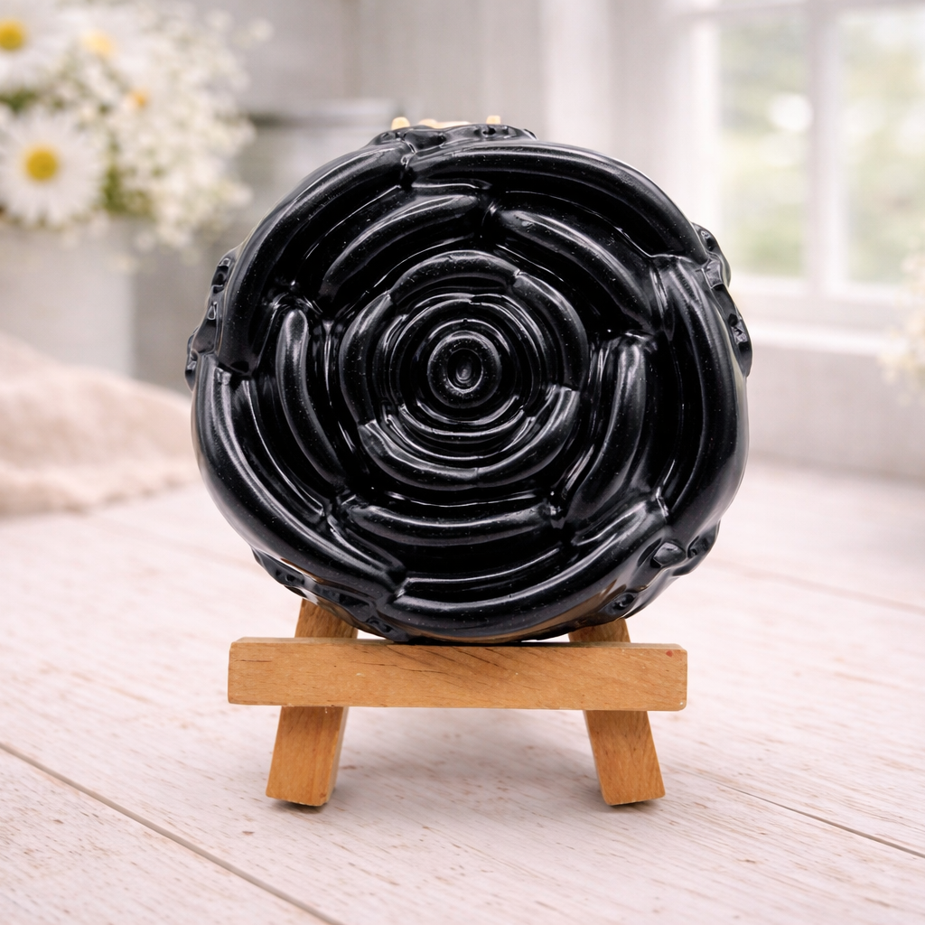 A handmade vegan soap, intricately sculpted in the shape of a large, blooming rose flower with layered petals, displayed on a small wooden easel against a bright win