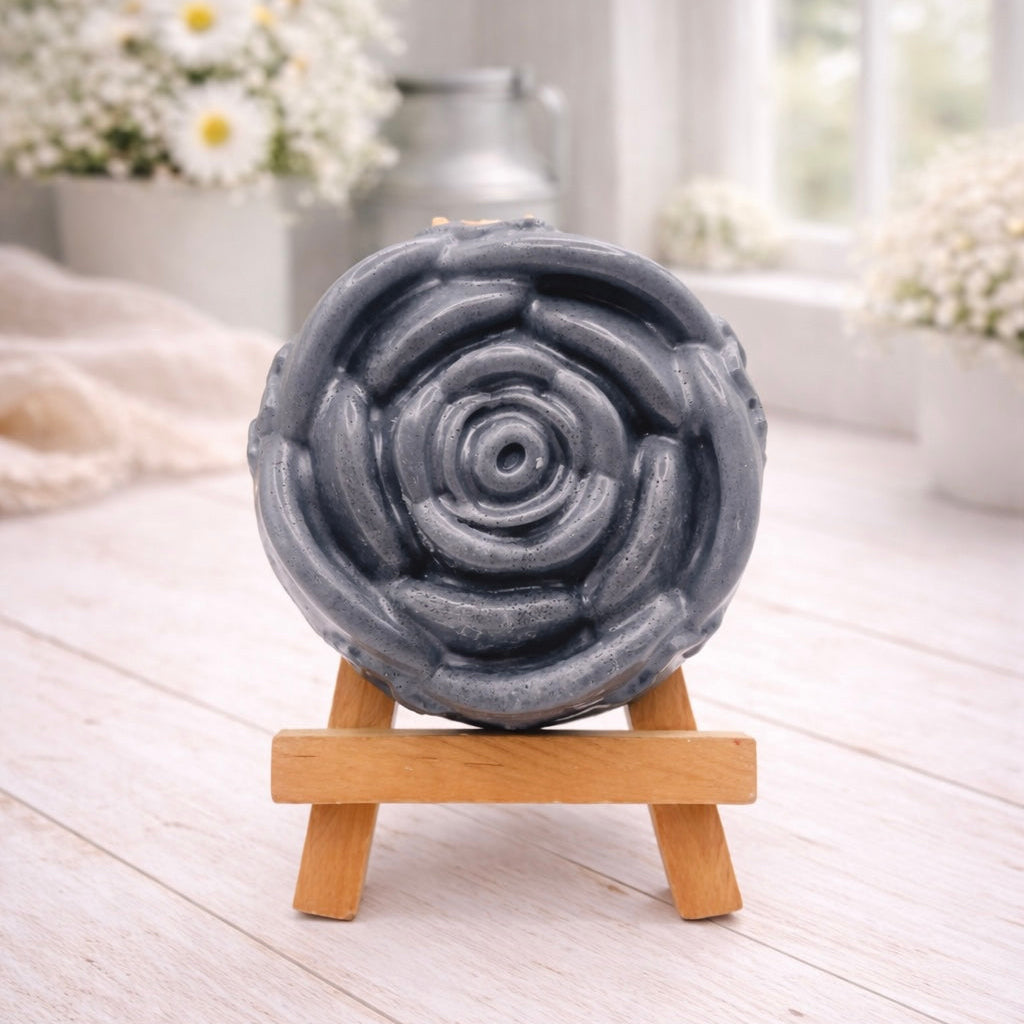 A handmade vegan soap, intricately sculpted in the shape of a large, blooming rose flower with layered petals, displayed on a small wooden easel against a bright win
