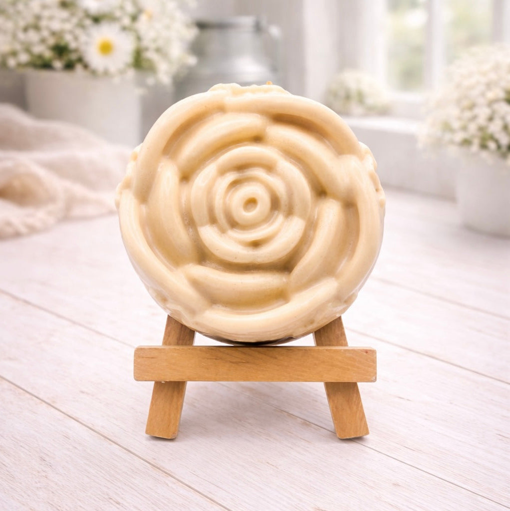 A handmade vegan soap, intricately sculpted in the shape of a large, blooming rose flower with layered petals, displayed on a small wooden easel against a bright win