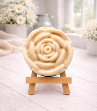 Load image into Gallery viewer, A handmade vegan soap, intricately sculpted in the shape of a large, blooming rose flower with layered petals, displayed on a small wooden easel against a bright win
