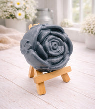 Load image into Gallery viewer, A handmade vegan soap, intricately sculpted in the shape of a large, blooming rose flower with layered petals, displayed on a small wooden easel against a bright win
