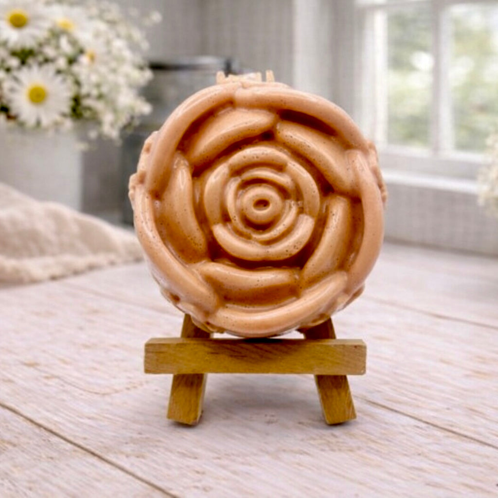 A handmade vegan soap, intricately sculpted in the shape of a large, blooming rose flower with layered petals, displayed on a small wooden easel against a bright win