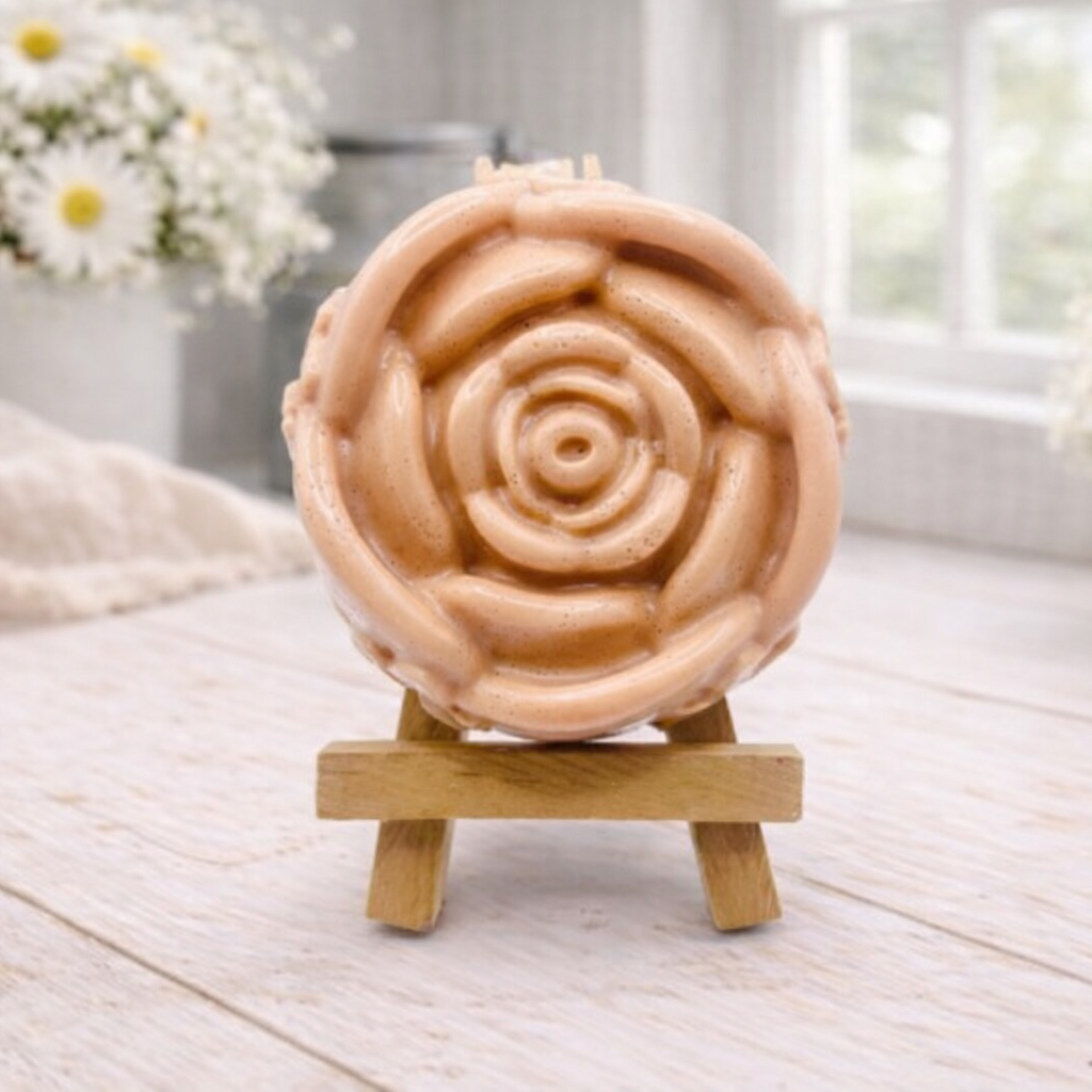 A handmade vegan soap, intricately sculpted in the shape of a large, blooming rose flower with layered petals, displayed on a small wooden easel against a bright win
