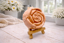 Load image into Gallery viewer, A handmade vegan soap, intricately sculpted in the shape of a large, blooming rose flower with layered petals, displayed on a small wooden easel against a bright win
