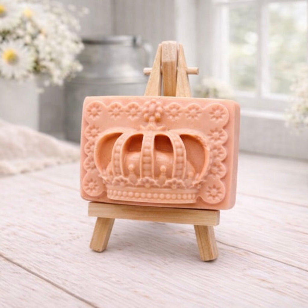 A rectangular handmade vegan soap in a soft tone, featuring a highly detailed relief of a royal crown with floral border accents, displayed on a miniature wooden easel in a bright farmhouse setting.