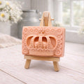 A rectangular handmade vegan soap in a soft tone, featuring a highly detailed relief of a royal crown with floral border accents, displayed on a miniature wooden easel in a bright farmhouse setting.