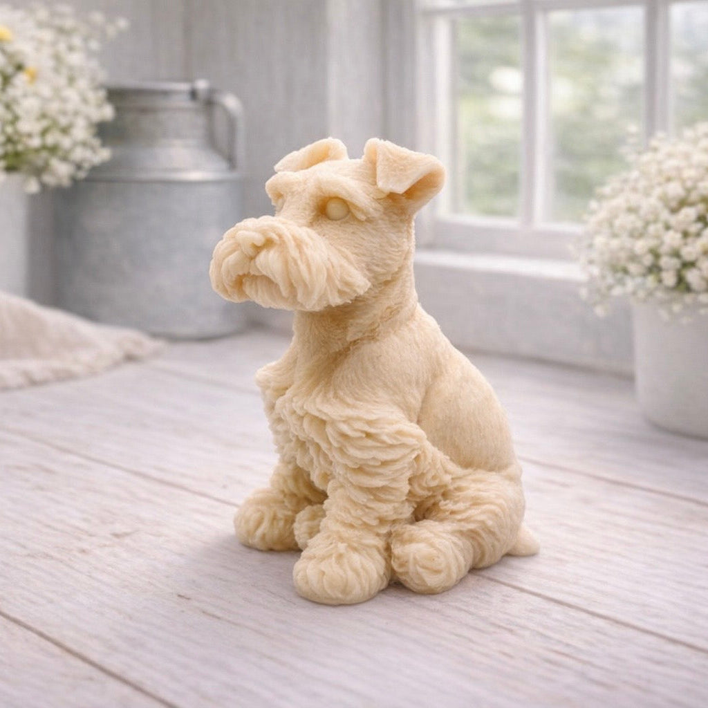 A detailed handmade vegan soap sculpted in the shape of a sitting Schnauzer dog with characteristic fluffy eyebrows and beard, in a natural colour, set against a bright window and white flowers. By soap Simple creations 