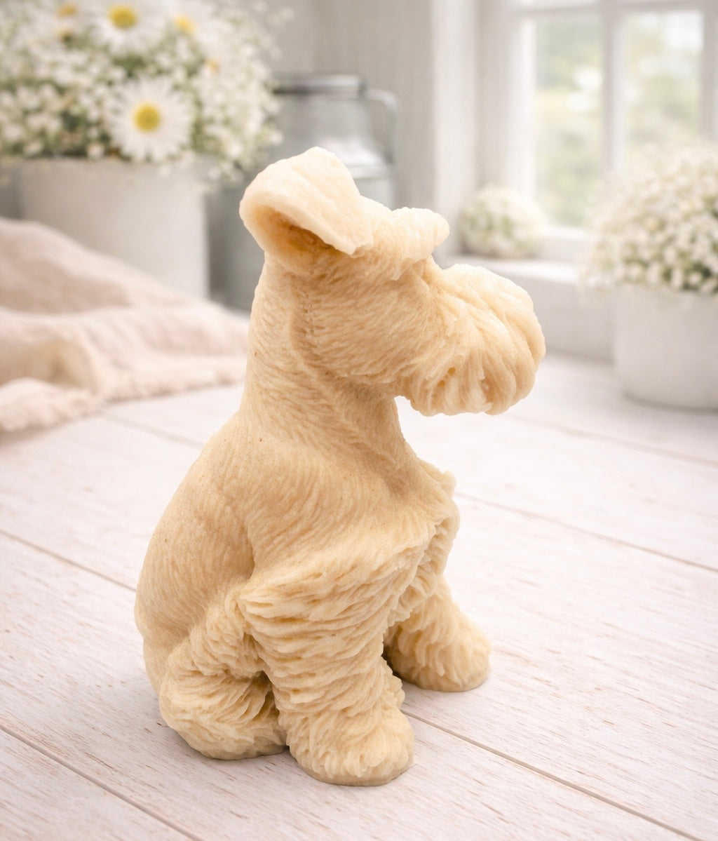 A detailed handmade vegan soap sculpted in the shape of a sitting Schnauzer dog with characteristic fluffy eyebrows and beard, in a natural colour, set against a bright window and white flowers. By soap Simple creations 