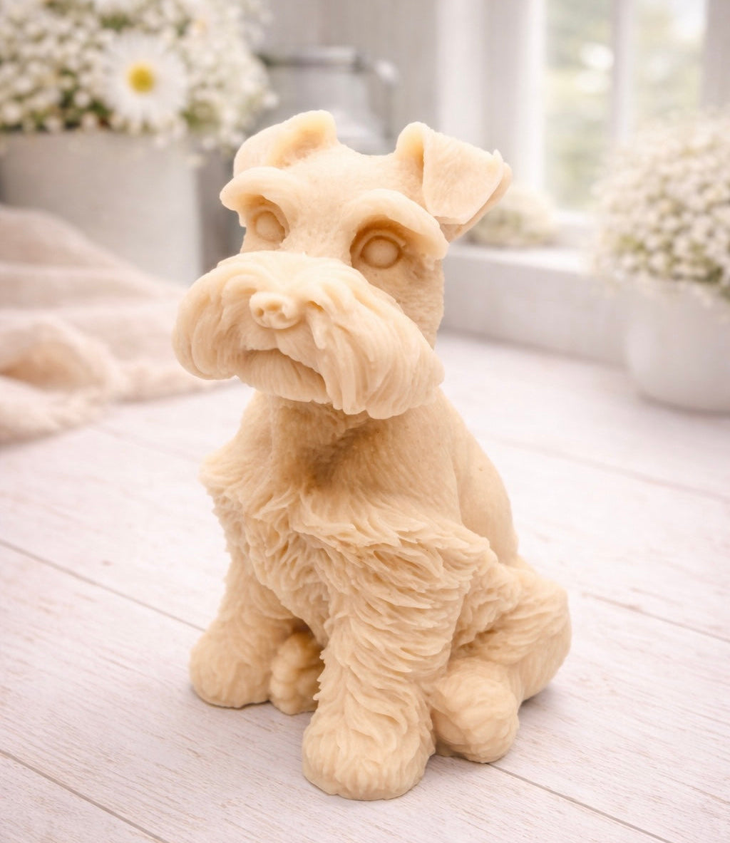 A detailed handmade vegan soap sculpted in the shape of a sitting Schnauzer dog with characteristic fluffy eyebrows and beard, in a natural colour, set against a bright window and white flowers. By soap Simple creations 