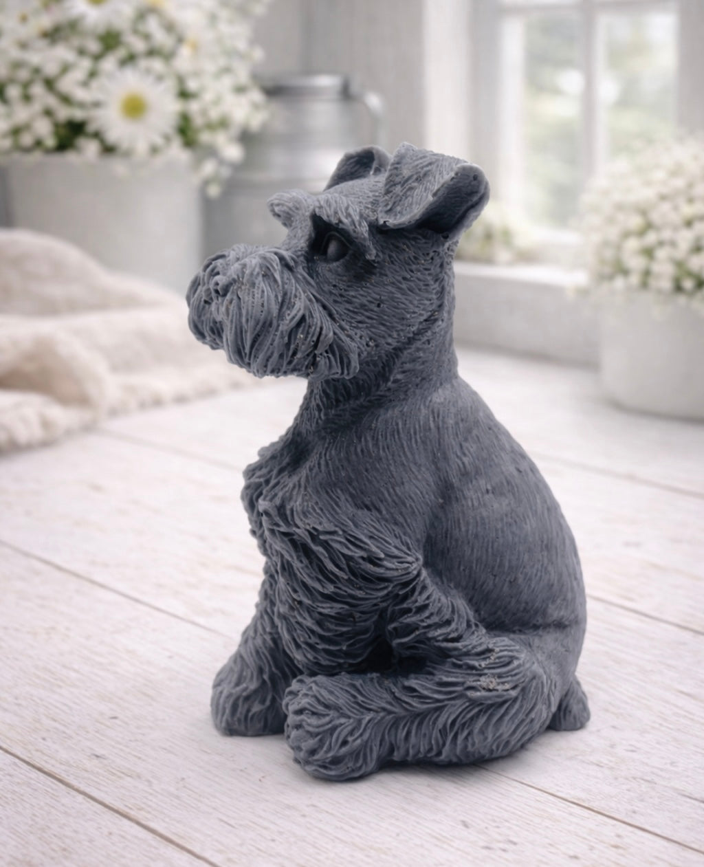 A detailed handmade vegan soap sculpted in the shape of a sitting Schnauzer dog with characteristic fluffy eyebrows and beard, in a natural colour, set against a bright window and white flowers. By soap Simple creations 