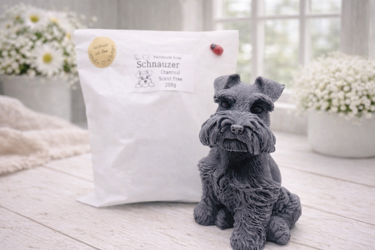 A detailed handmade vegan soap sculpted in the shape of a sitting Schnauzer dog with characteristic fluffy eyebrows and beard, in a natural colour, set against a bright window and white flowers. By soap Simple creations 