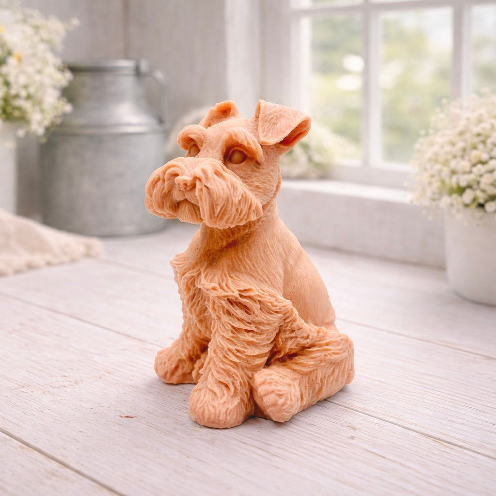 A detailed handmade vegan soap sculpted in the shape of a sitting Schnauzer dog with characteristic fluffy eyebrows and beard, in a natural colour, set against a bright window and white flowers. By soap Simple creations 