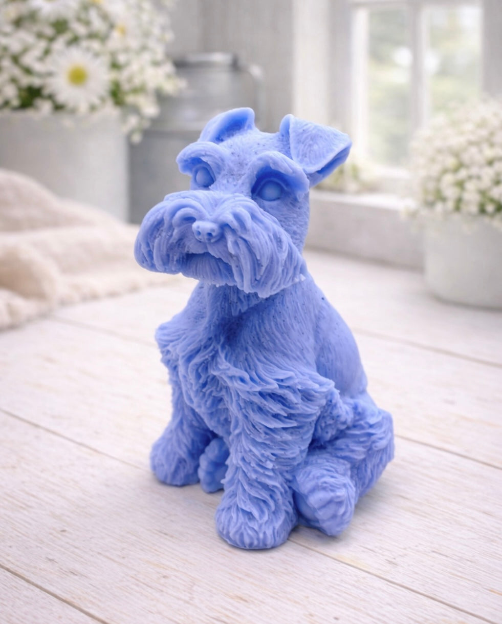 A detailed handmade vegan soap sculpted in the shape of a sitting Schnauzer dog with characteristic fluffy eyebrows and beard, in a natural colour, set against a bright window and white flowers. By soap Simple creations 