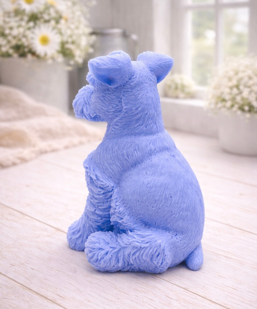 A detailed handmade vegan soap sculpted in the shape of a sitting Schnauzer dog with characteristic fluffy eyebrows and beard, in a natural colour, set against a bright window and white flowers. By soap Simple creations 