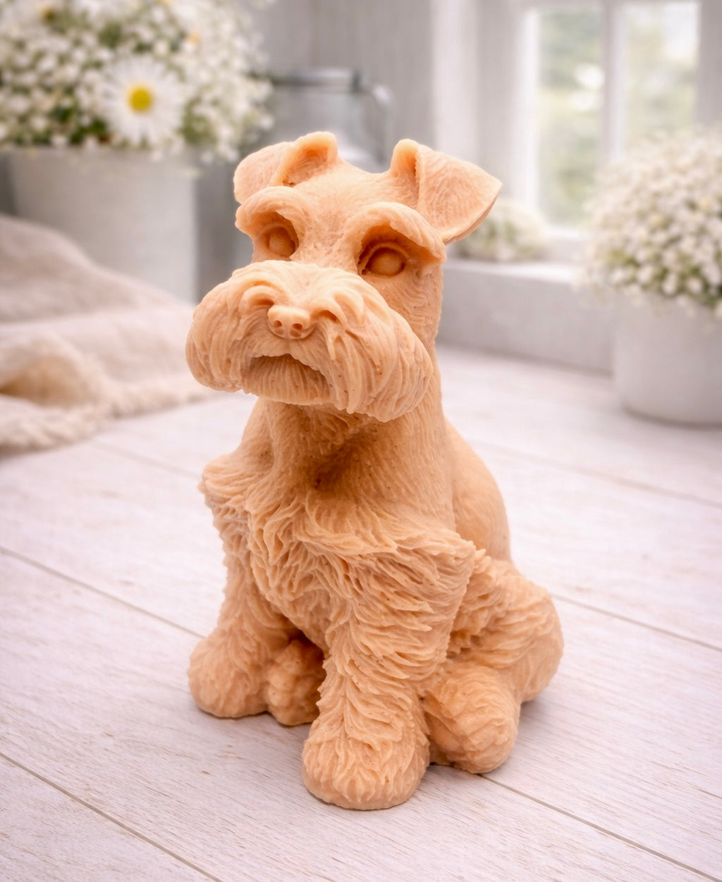 A detailed handmade vegan soap sculpted in the shape of a sitting Schnauzer dog with characteristic fluffy eyebrows and beard, in a natural colour, set against a bright window and white flowers. By soap Simple creations 