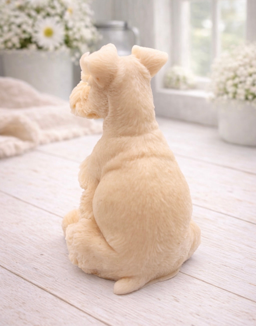 A detailed handmade vegan soap sculpted in the shape of a sitting Schnauzer dog with characteristic fluffy eyebrows and beard, in a natural colour, set against a bright window and white flowers. By soap Simple creations 
