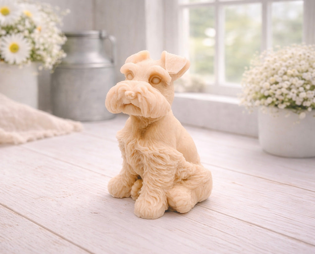 A detailed handmade vegan soap sculpted in the shape of a sitting Schnauzer dog with characteristic fluffy eyebrows and beard, in a natural colour, set against a bright window and white flowers. By soap Simple creations 