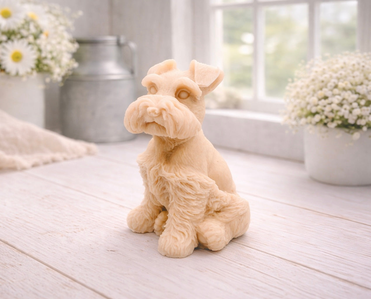 A detailed handmade vegan soap sculpted in the shape of a sitting Schnauzer dog with characteristic fluffy eyebrows and beard, in a natural colour, set against a bright window and white flowers. By soap Simple creations 
