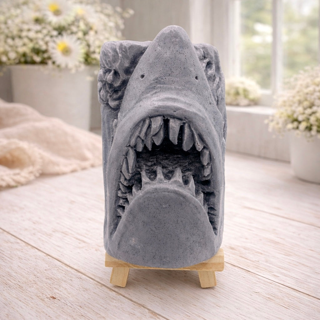 A 3D handmade vegan soap sculpted into a realistic great white shark head with its jaws wide open to reveal multiple rows of sharp teeth, displayed on a miniature wooden easel in a bright farmhouse setting.