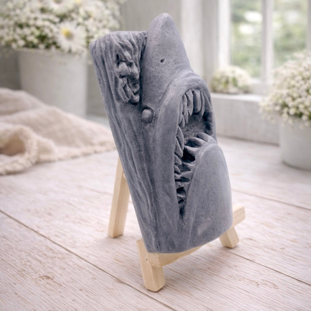 A 3D handmade vegan soap sculpted into a realistic great white shark head with its jaws wide open to reveal multiple rows of sharp teeth, displayed on a miniature wooden easel in a bright farmhouse setting.