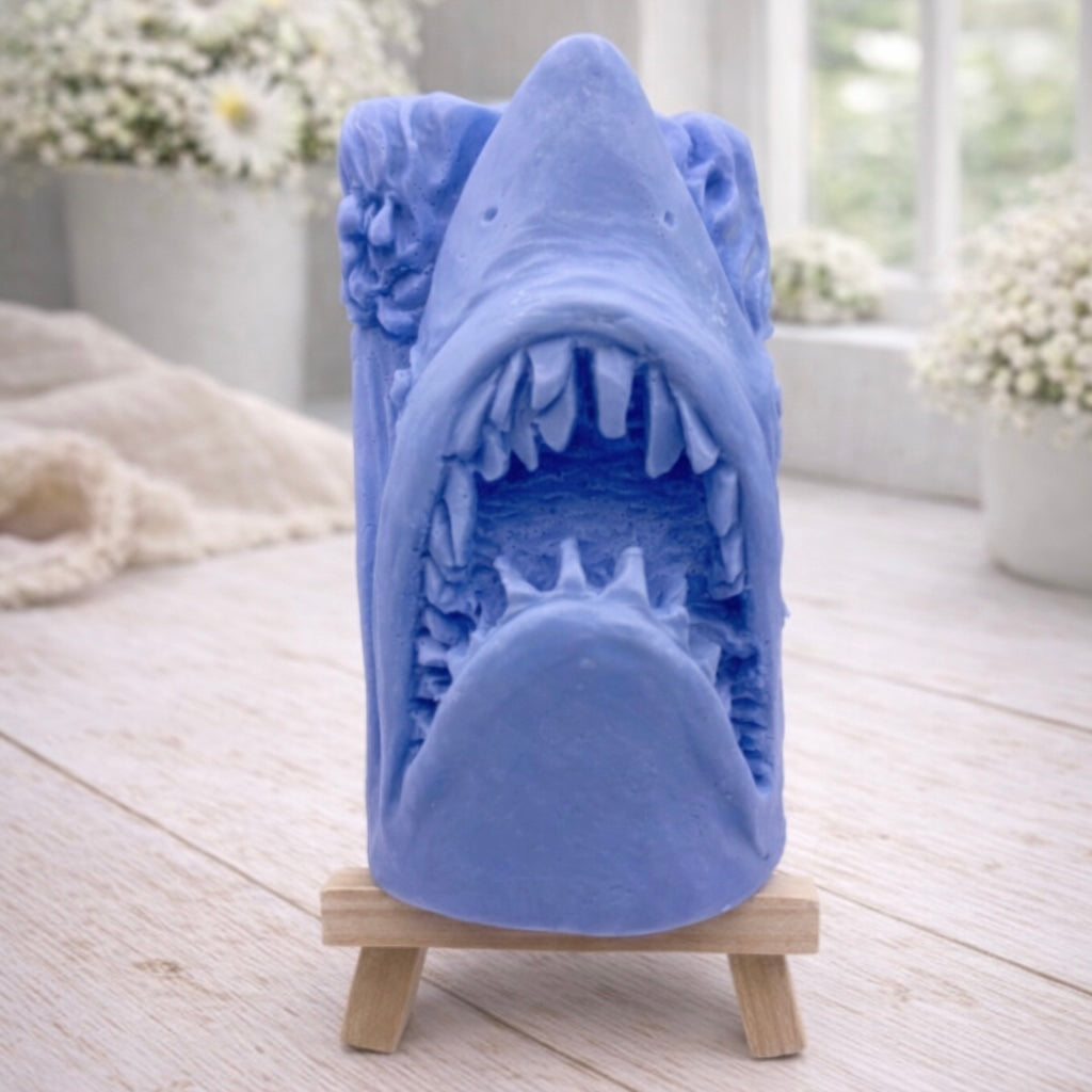A 3D handmade vegan soap sculpted into a realistic great white shark head with its jaws wide open to reveal multiple rows of sharp teeth, displayed on a miniature wooden easel in a bright farmhouse setting.