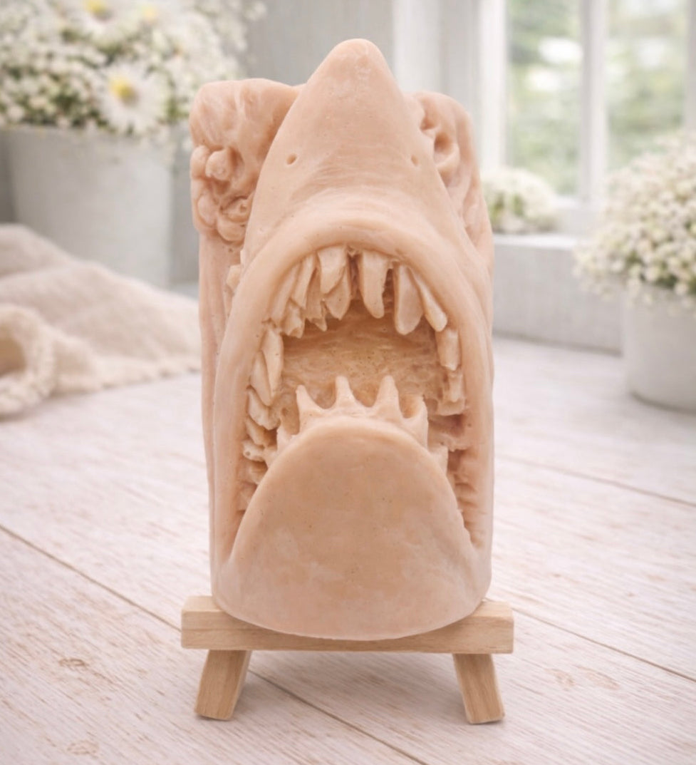 A 3D handmade vegan soap sculpted into a realistic great white shark head with its jaws wide open to reveal multiple rows of sharp teeth, displayed on a miniature wooden easel in a bright farmhouse setting.