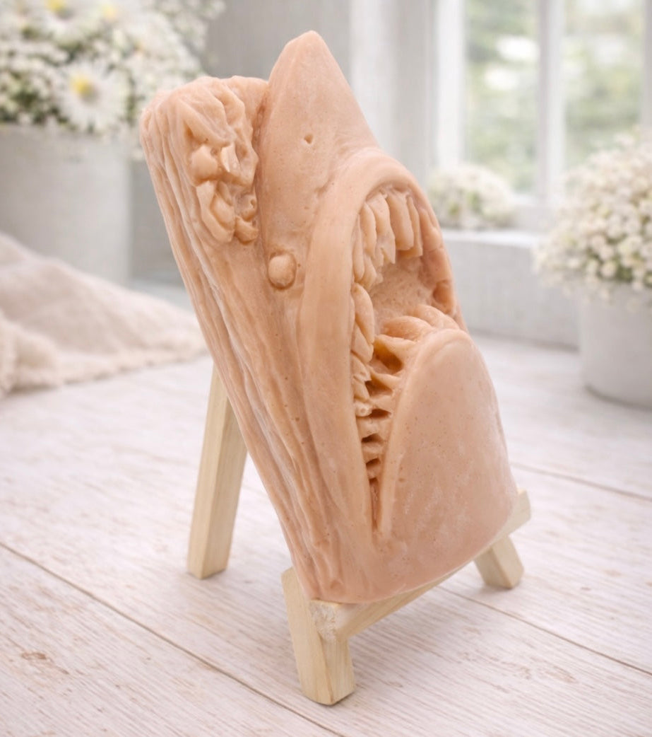 A 3D handmade vegan soap sculpted into a realistic great white shark head with its jaws wide open to reveal multiple rows of sharp teeth, displayed on a miniature wooden easel in a bright farmhouse setting.