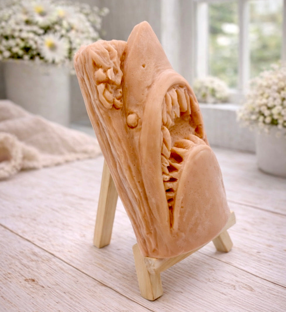 A 3D handmade vegan soap sculpted into a realistic great white shark head with its jaws wide open to reveal multiple rows of sharp teeth, displayed on a miniature wooden easel in a bright farmhouse setting.