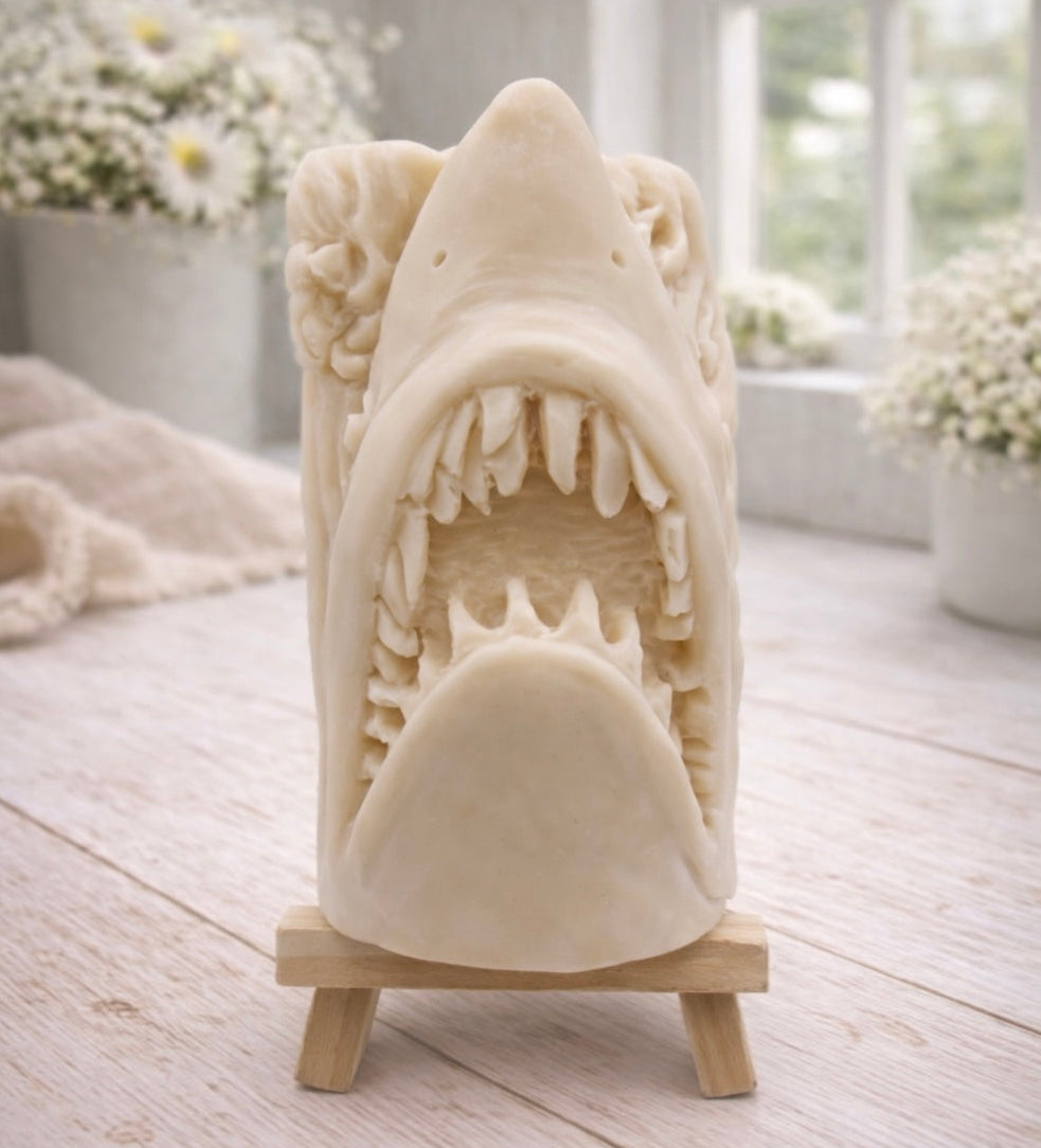 A 3D handmade vegan soap sculpted into a realistic great white shark head with its jaws wide open to reveal multiple rows of sharp teeth, displayed on a miniature wooden easel in a bright farmhouse setting.