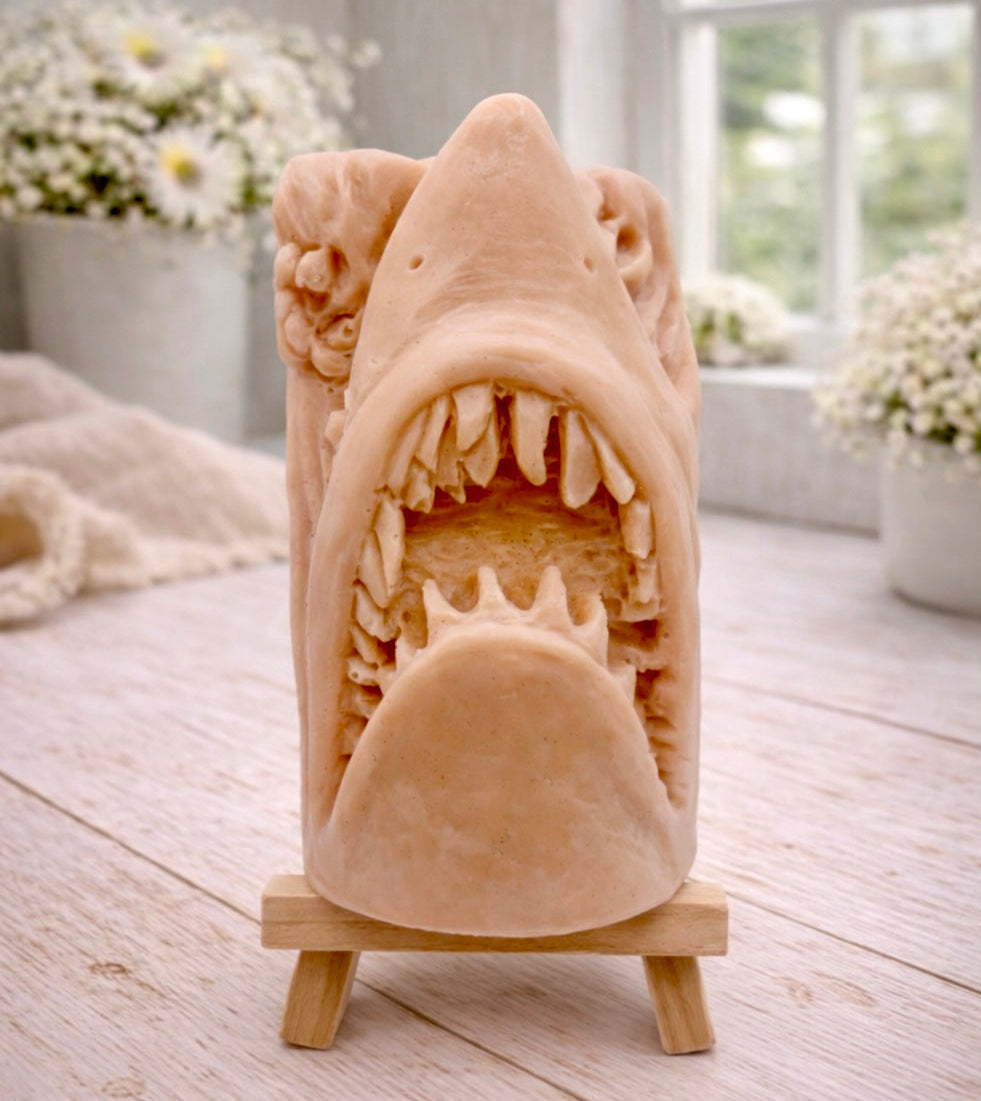 A 3D handmade vegan soap sculpted into a realistic great white shark head with its jaws wide open to reveal multiple rows of sharp teeth, displayed on a miniature wooden easel in a bright farmhouse setting.