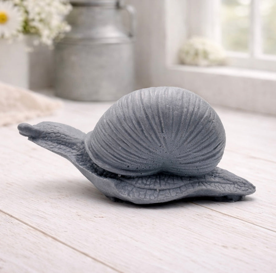 A highly detailed handmade vegan soap gift, sculpted in the shape of a realistic snail with a prominent spiralled shell and a textured, leathery body, displayed on a light wooden surface. Handmade in Blackpool. 