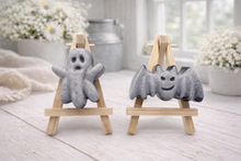 Load image into Gallery viewer, A collection of six handmade vegan Halloween soaps in a mottled grey and black charcoal finish, featuring a smiling bat, three different carved pumpkins (one wearing a witch's hat), a ghost, and a skull, arranged on and around a small wooden easel.
