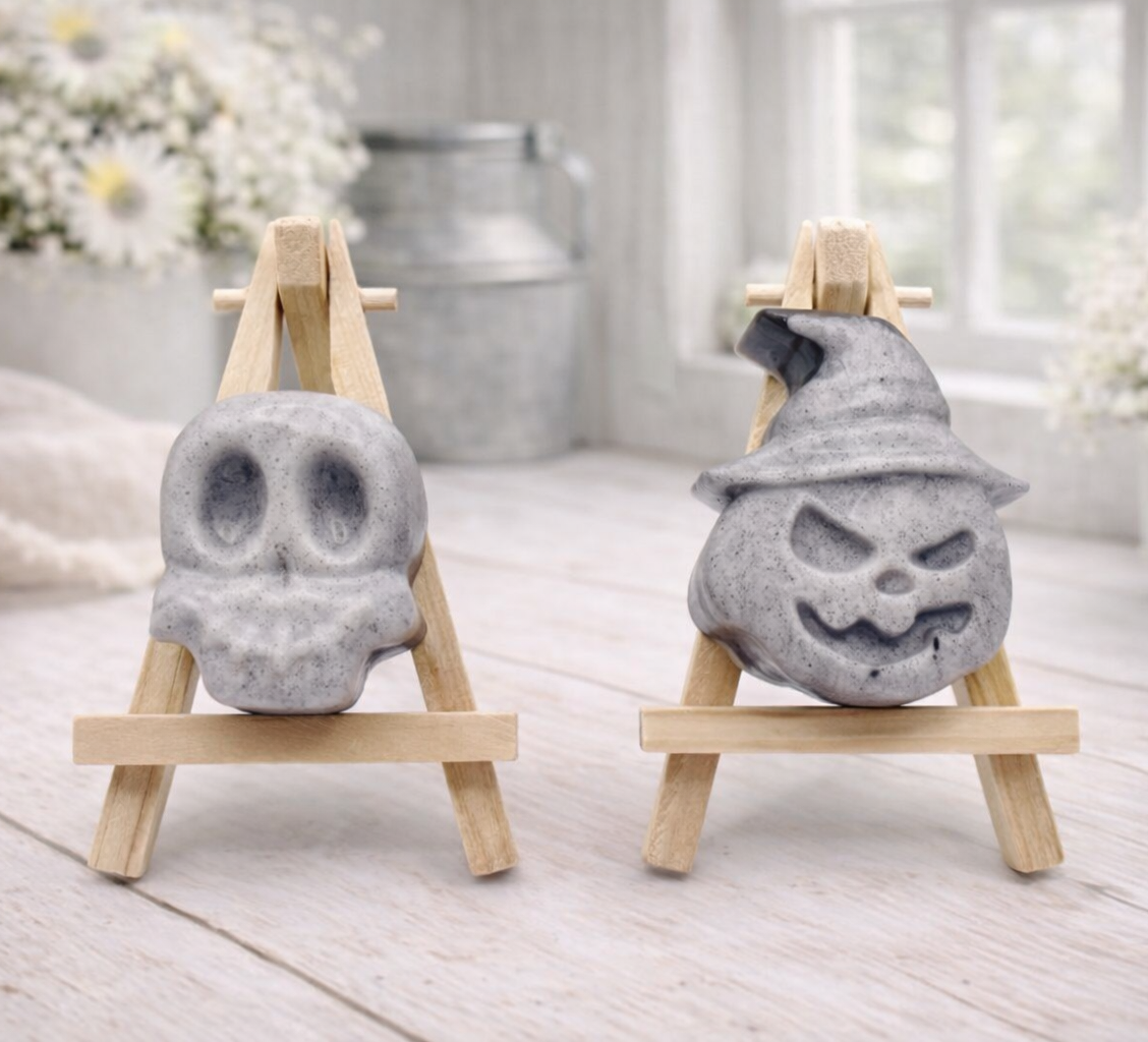 Two vegan soap sculptures of a skull and a jack-o'-lantern on small wooden stands in a bright room.