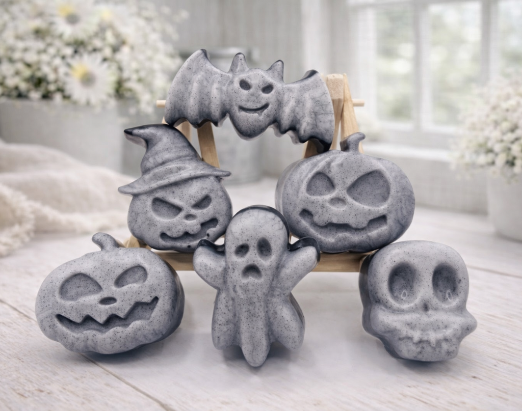 A collection of six handmade vegan Halloween soaps in a mottled grey and black charcoal finish, featuring a smiling bat, three different carved pumpkins (one wearing a witch's hat), a ghost, and a skull, arranged on and around a small wooden easel.