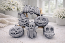 Load image into Gallery viewer, A collection of six handmade vegan Halloween soaps in a mottled grey and black charcoal finish, featuring a smiling bat, three different carved pumpkins (one wearing a witch's hat), a ghost, and a skull, arranged on and around a small wooden easel.
