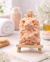 Load image into Gallery viewer, Handmade pig-shaped soap bar displayed on a small wooden easel, surrounded by spa items like bath salts, towel, brush, and oil bottle in a bright, calming bathroom setting.
