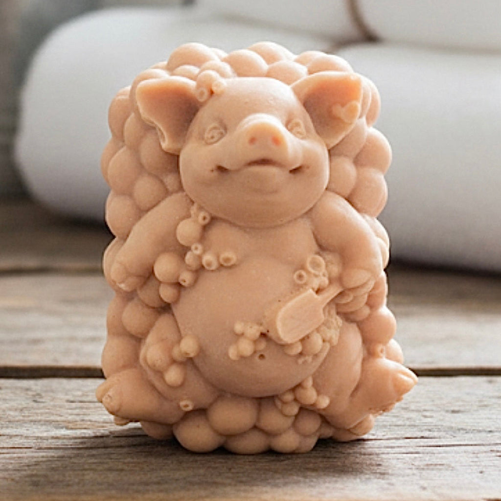 A detailed handmade vegan soap, sculpted as a cheerful pig sitting in a mound of bubbles and holding a scrubbing brush, displayed on a miniature wooden easel against a soft farmhouse window background 